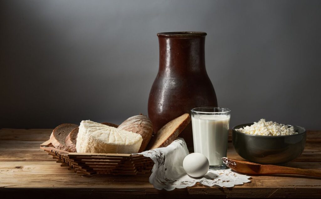 A rustic still life featuring bread, cheese, egg, milk, and cottage cheese on a wooden table.